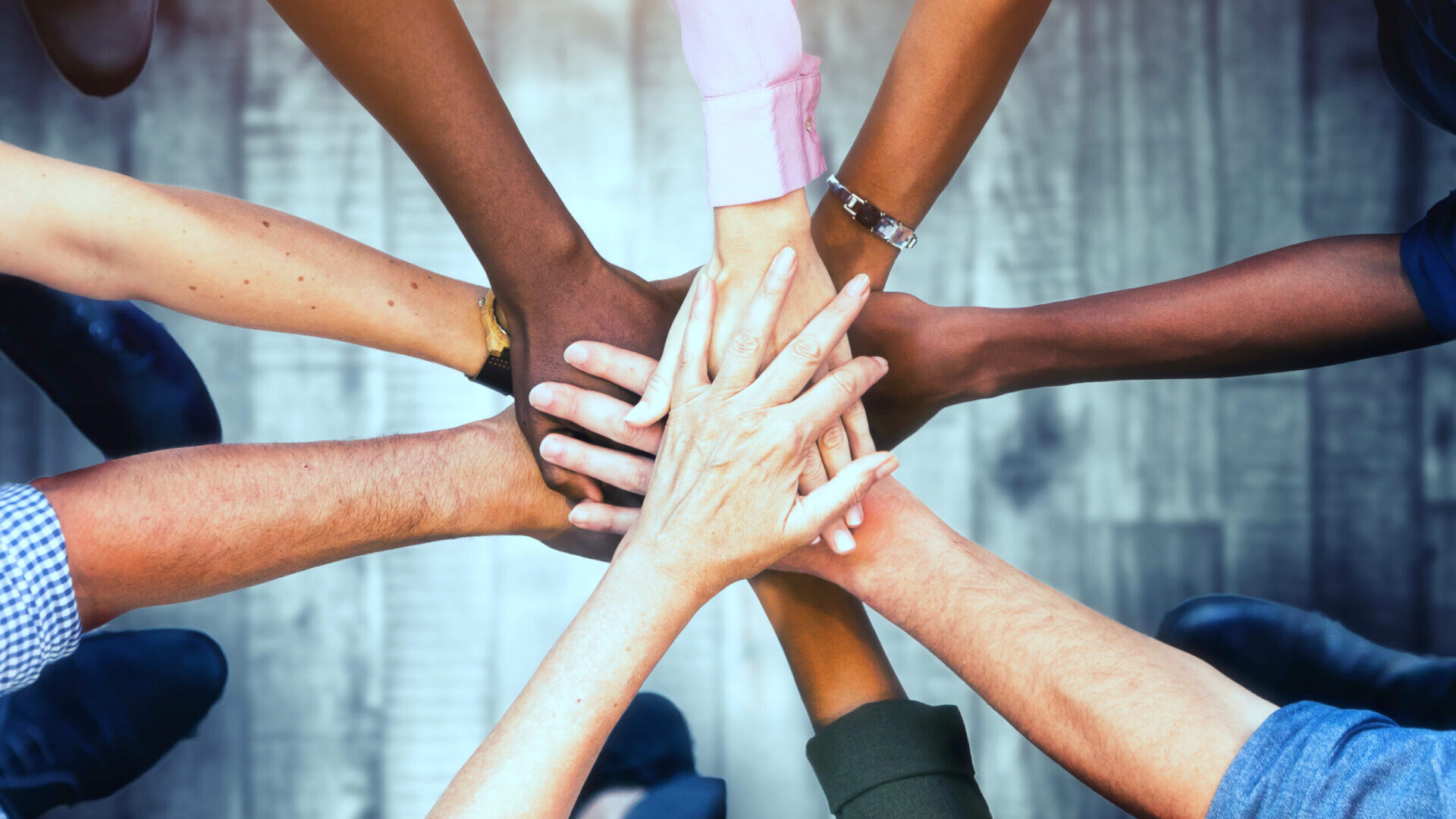 Overhead view of arms and hands reaching to a central point, like spokes on a wheel. The hands, representing various races or ethic groups, are all laid one on top of the other.